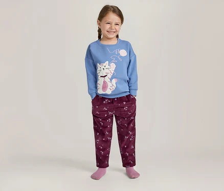 Lächelndes Mädchen mit Zöpfen, das ein blaues Langarmshirt mit Katzenmotiv und eine weinrote Hose mit Blumenmuster trägt.