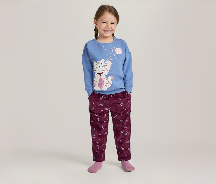 Lächelndes Mädchen mit Zöpfen, das ein blaues Langarmshirt mit Katzenmotiv und eine weinrote Hose mit Blumenmuster trägt.