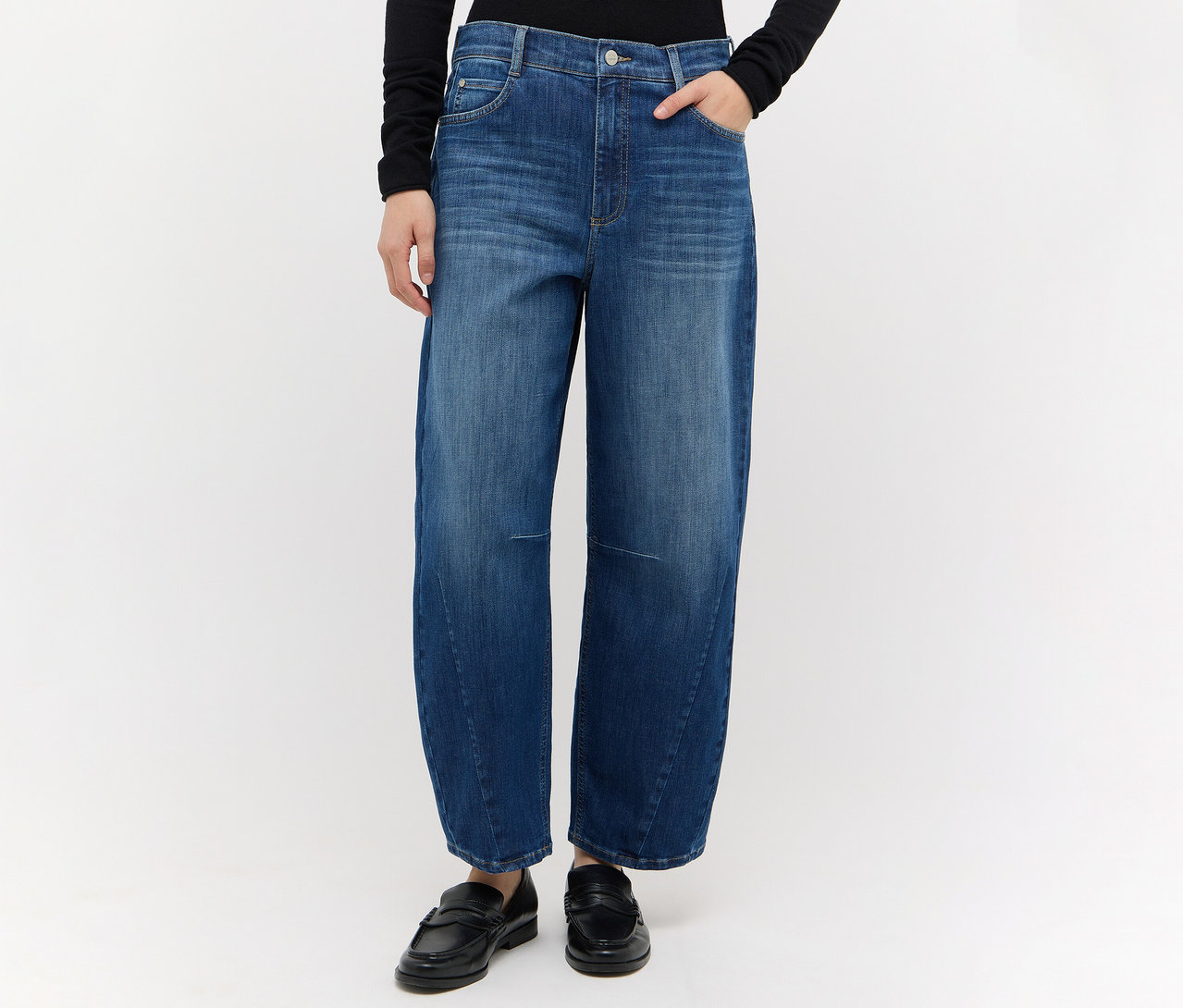 Person trägt eine weit geschnittene blaue Angels Denim Elly Jeans, ein schwarzes Langarmshirt und schwarze Loafer, eine Hand steckt in der Tasche.