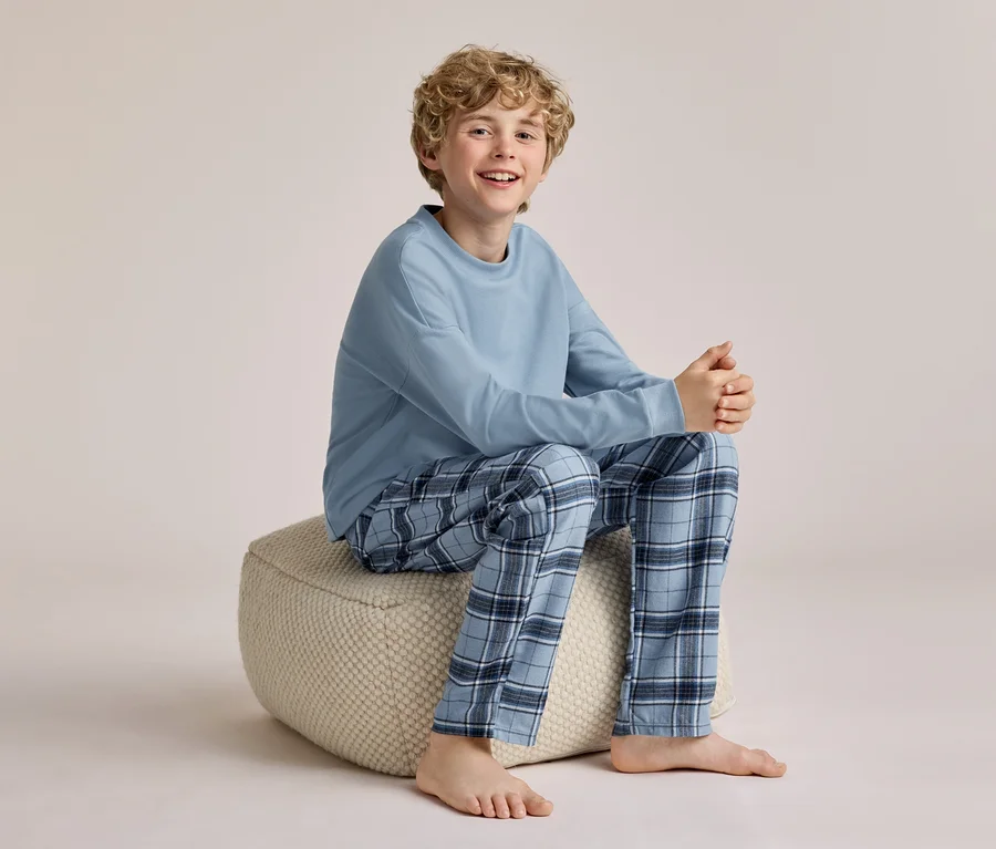 Ein lächelnder Junge mit lockigem blondem Haar sitzt barfuß auf einem hellen Pouf und trägt ein hellblaues Langarmshirt und blau karierte Pyjamahosen.