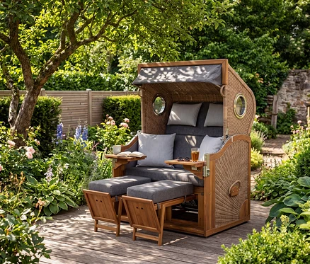 Ein XL deVries Strandkorb aus Akazienholz und Korbgeflecht mit grauen Polstern steht auf einer Holzterrasse in einem Garten. Ausziehbare Fußstützen und Beistelltische mit Buch, Tasse, Getränk und Sonnenbrille.