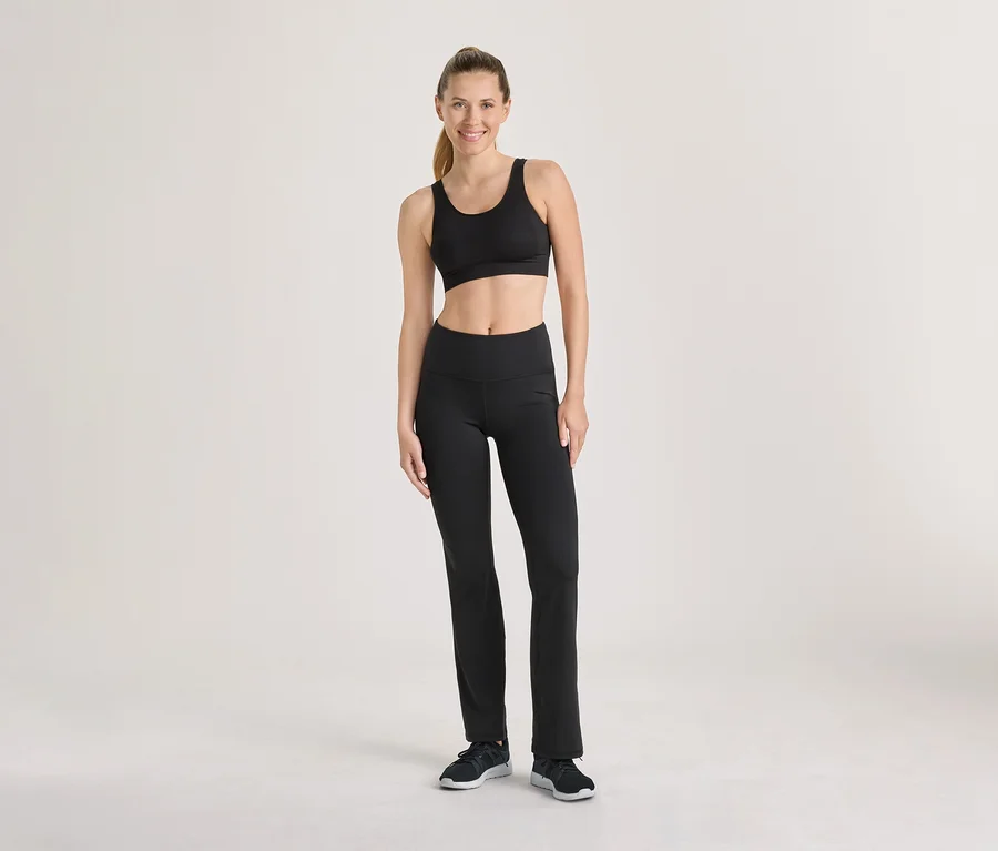 Lächelnde Frau steht in schwarzem Sport-BH, schwarzer Sporthose und schwarzen Turnschuhen.