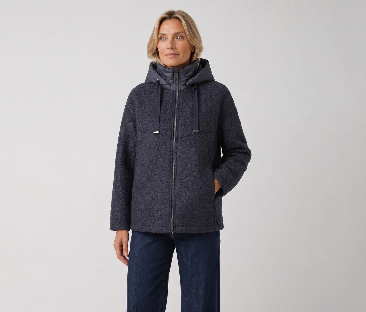 Blonde Frau in einer dunkelblauen, strukturierten Kapuzenjacke mit Reißverschluss und Jeans, die Hand in der Tasche.