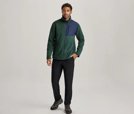 Mann steht in einer grün-marineblauen Fleecejacke, schwarzen Hosen und dunklen Wanderschuhen, die linke Hand in der Tasche.