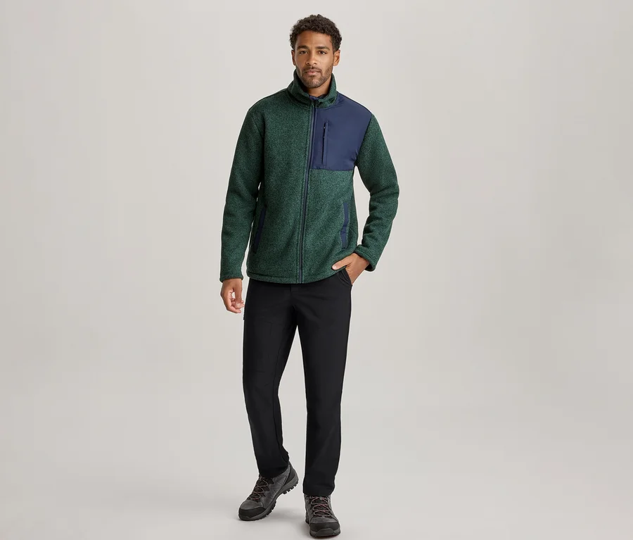 Mann steht in einer grün-marineblauen Fleecejacke, schwarzen Hosen und dunklen Wanderschuhen, die linke Hand in der Tasche.