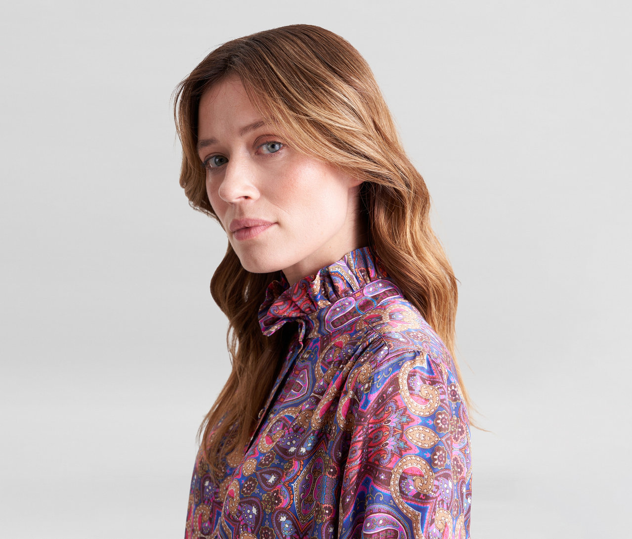 Frau mit langen braunen Haaren trägt eine bunte Paisley-Bluse mit Rüschenkragen und blickt in die Kamera.