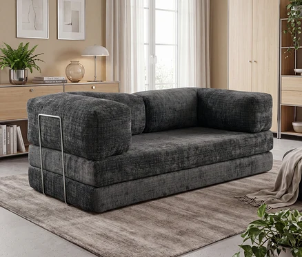 Dunkelgraues Modulsofa mit dicken Polstern und einer Armlehnenstütze aus Metall links, auf einem strukturierten grauen Teppich in einem Wohnzimmer. Im Hintergrund sind Holzmöbel und ein großes Fenster zu sehen.