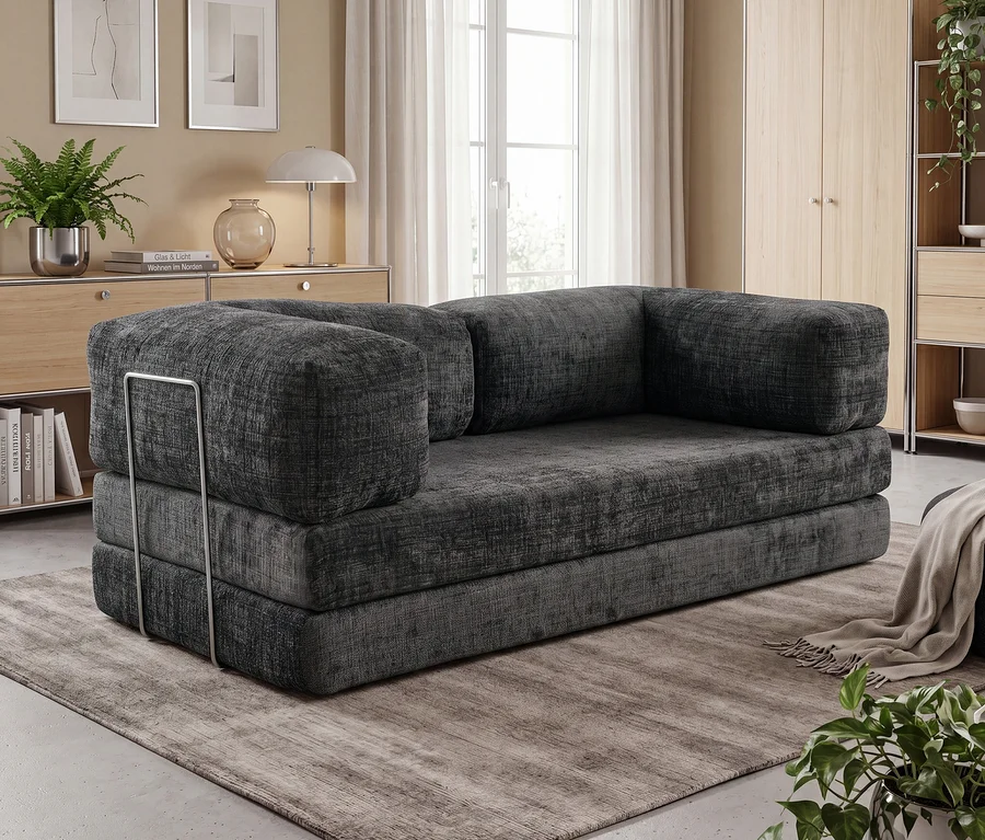 Dunkelgraues Modulsofa mit dicken Polstern und einer Armlehnenstütze aus Metall links, auf einem strukturierten grauen Teppich in einem Wohnzimmer. Im Hintergrund sind Holzmöbel und ein großes Fenster zu sehen.