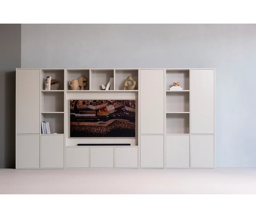 Helles, modulares Mediamöbel mit offenen Regalen und geschlossenen Schränken. Ein Fernseher mit einer Luftaufnahme einer Küstenstadt und eine Soundbar sind in der Mitte platziert. Die Regale sind mit Vasen, Skulpturen und Büchern dekoriert.