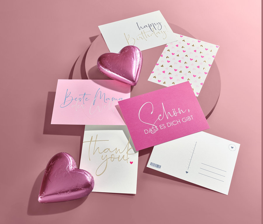 Draufsicht von Karten mit Schriftzügen und zwei rosa Herzen auf rosafarbenem Hintergrund.