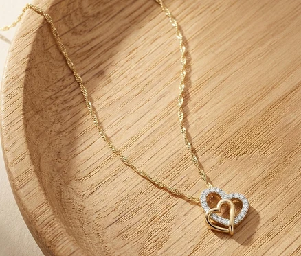 Goldene Amor Halskette mit gedrehter Kette und einem Anhänger aus zwei ineinandergreifenden Herzen, eines davon mit weißen Steinen besetzt, auf hellem Holz.