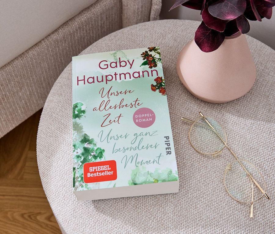 Auf einem beigen Hocker liegen der Doppelroman »Unsere allerbeste Zeit« und »Unser ganz besonderer Moment«, eine goldene Brille und eine Vase mit einer Pflanze.