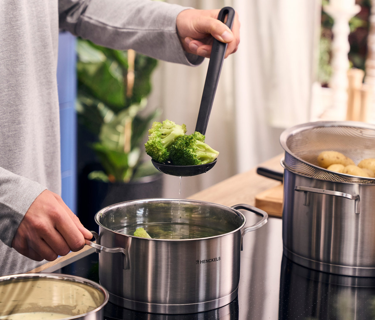 Eine Person hebt mit einem schwarzen Schaumlöffel Brokkoli aus einem Topf des Henckels by ZWILLING »Pulse« Kochgeschirrsets. Daneben steht ein weiterer Topf mit Kartoffeln.