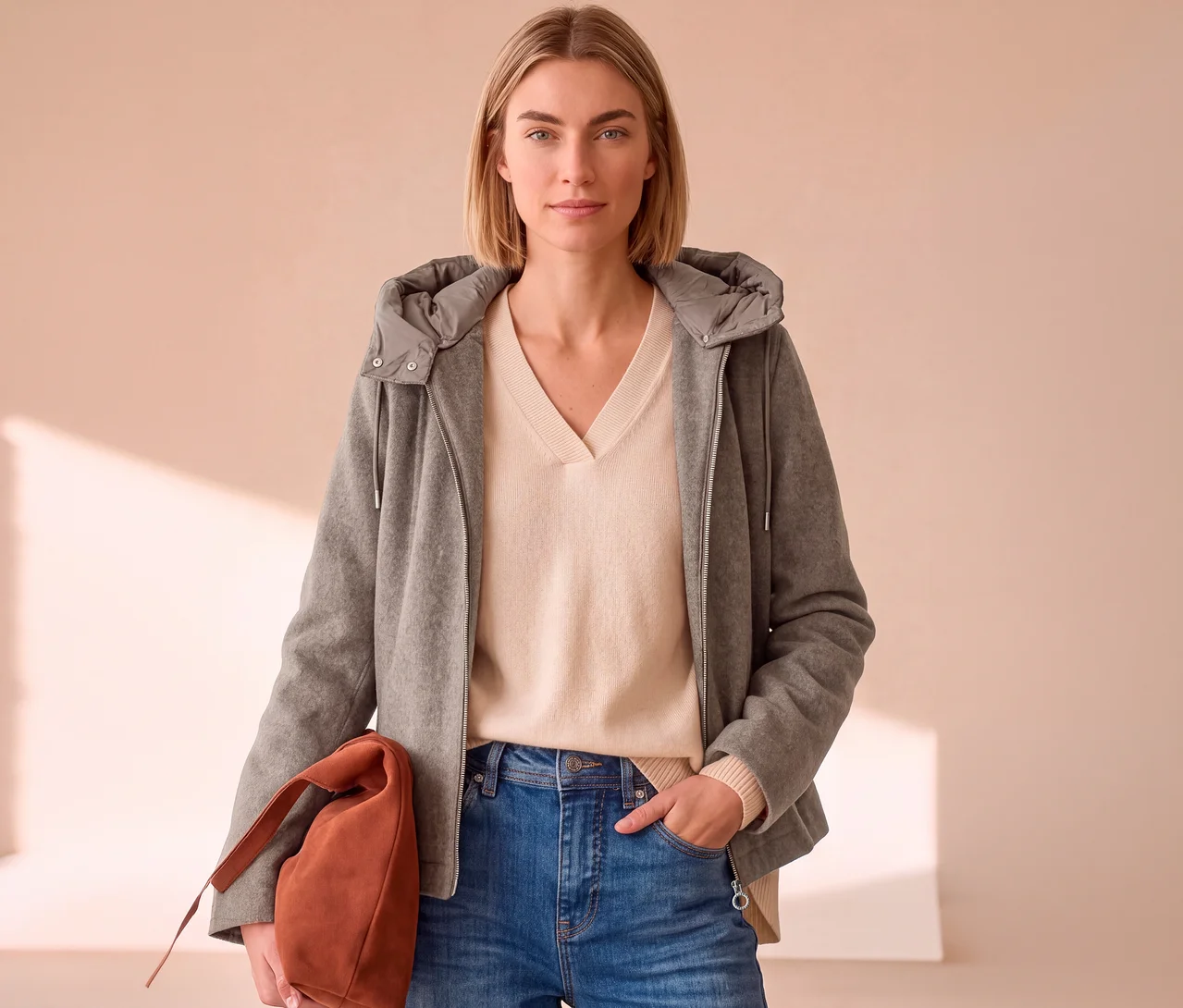 Eine Frau mit blonden Haaren trägt eine offene graue Street One Übergangsjacke über einem hellbeigen V-Pullover und blauen Jeans, eine braune Handtasche haltend.