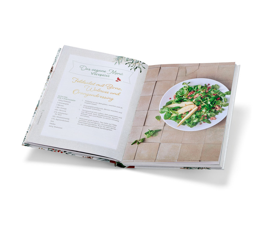 Aufgeschlagenes Kochbuch »Christmas is a feeling« mit Rezept für Feldsalat mit Birne und Walnussdressing und fertigem Salat auf einem Teller.