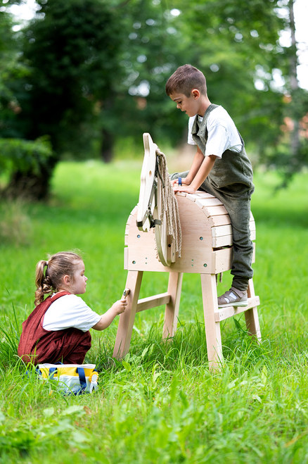Ein Junge sitzt auf einem Holzpferd, ein Mädchen steht daneben und spielt damit. Am Pferd ist ein Zaumzeug befestigt. Im Gras steht eine Tasche mit einem small foot Spielzeug-Pferdepflege-Set mit Tasche.