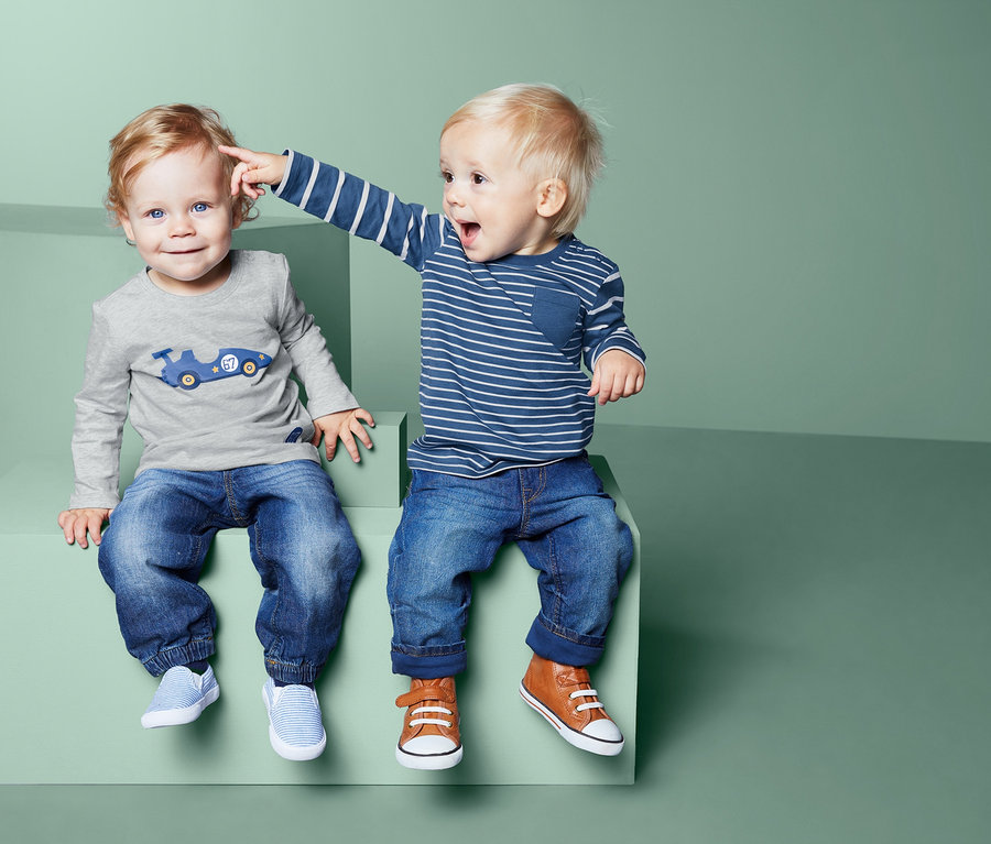 Zwei Jungen sitzen auf grünen Blöcken, einer tippt dem anderen an die Stirn. Beide tragen Jeans und 2 Langarmshirts.