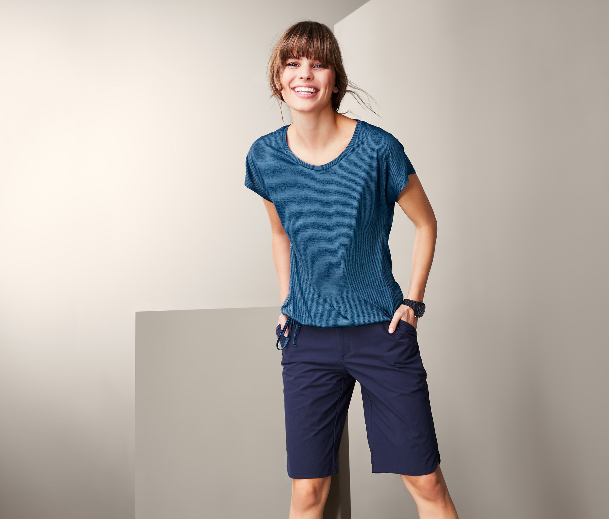 Eine lächelnde Frau posiert in einem blauen Funktionsshirt und einer dunkelblauen Shorts.