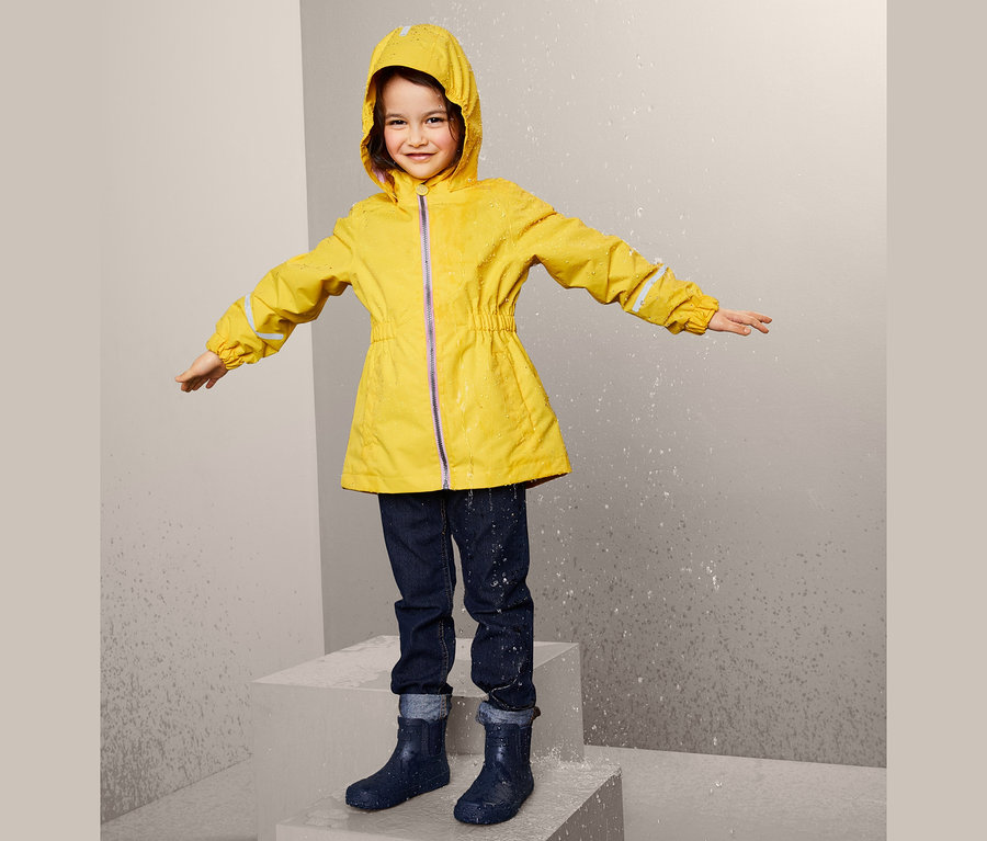 Ein Mädchen steht im Regen und trägt einen gelben Regenmantel aus recyceltem Material, Jeans und Gummistiefel.