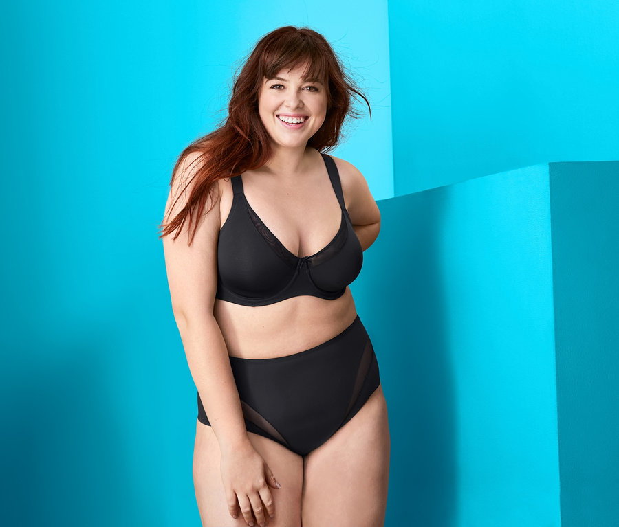 Frau trägt einen schwarzen Minimizer-BH und schwarze Slips vor einem Hintergrund mit blauen Formen.