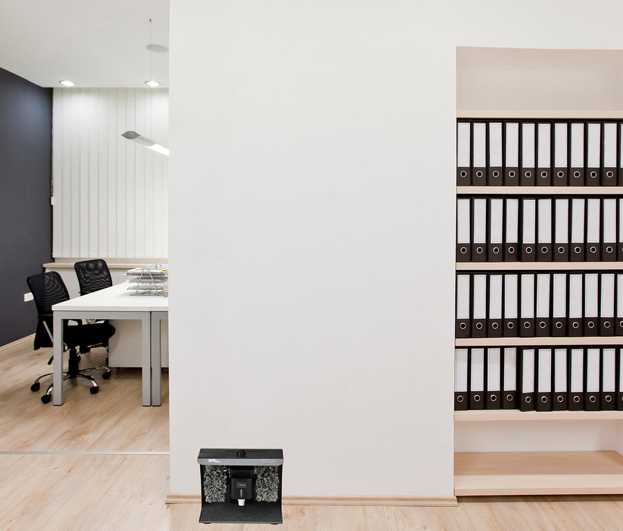 Im Vordergrund befindet sich eine Trebs Schuhputzmaschine. Im Hintergrund befindet sich ein Büro mit einem weißen Tisch, schwarzen Stühlen und einem Schrank voller Ordner.