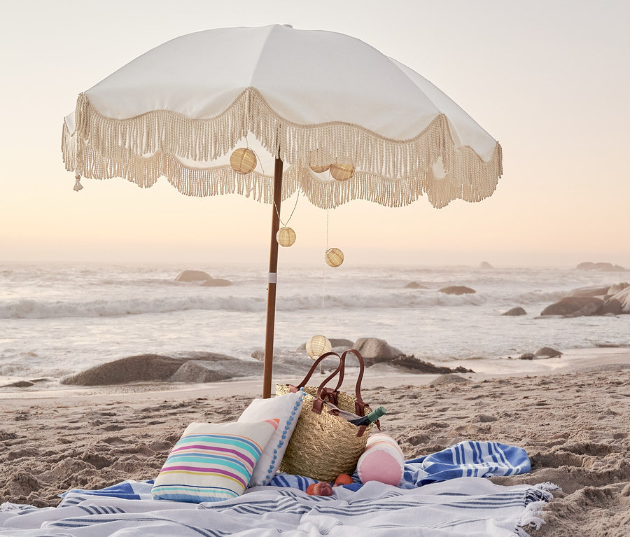 Strand mit einem Strand-Sonnenschirm, einer LED-Lichterkette, einer Korbtasche, einem Outdoor-Kissen mit Tasseln, einer Outdoor-Kissenrolle und einem Outdoor-Kissen.