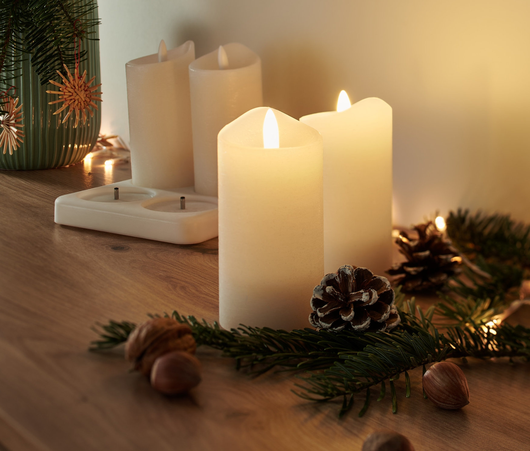 Vier LED-Echtwachskerzen mit Akku stehen auf einem Holztisch neben einer Vase mit Tannenzweigen, Zapfen und Nüssen.