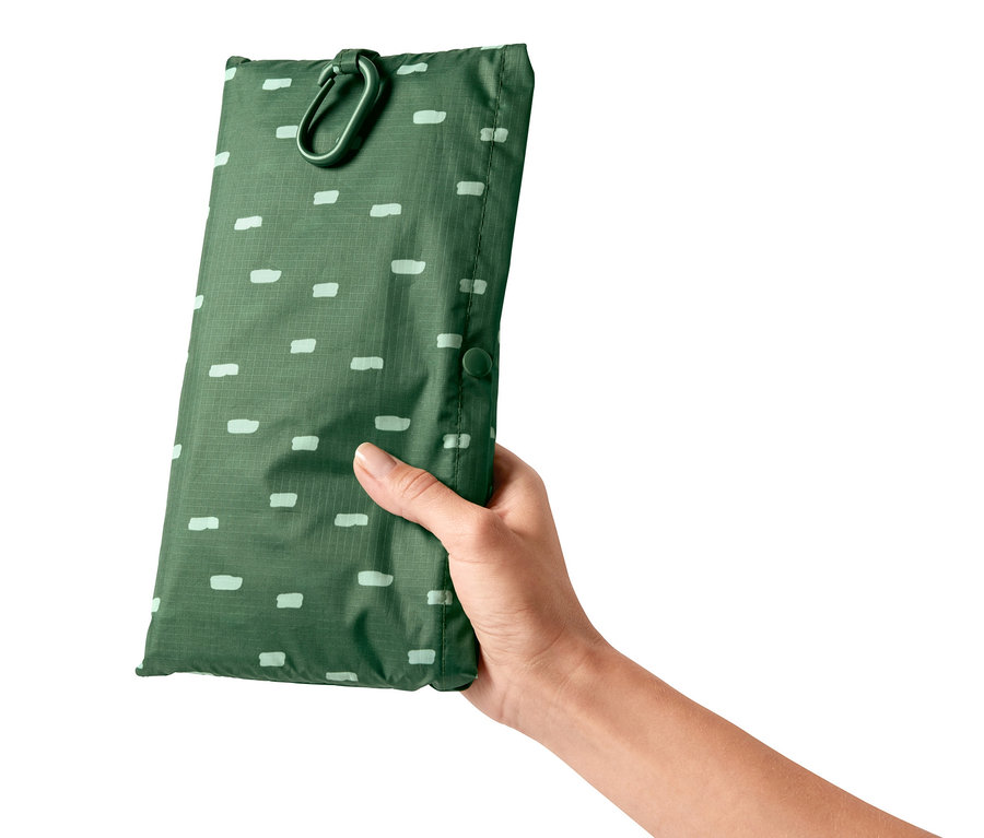 Eine Hand hält eine grüne 2-in-1-Picknickdecke mit hellen Rechtecken und Karabiner.