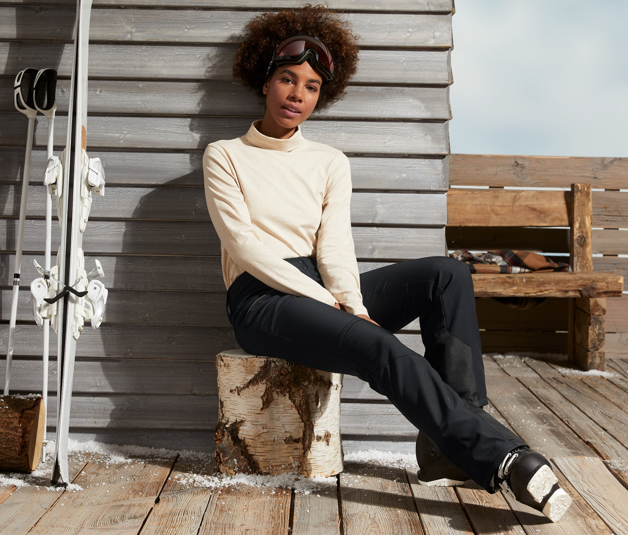 Frau sitzt auf Holzscheit in schwarzer Jetskihose und Skischuhen.