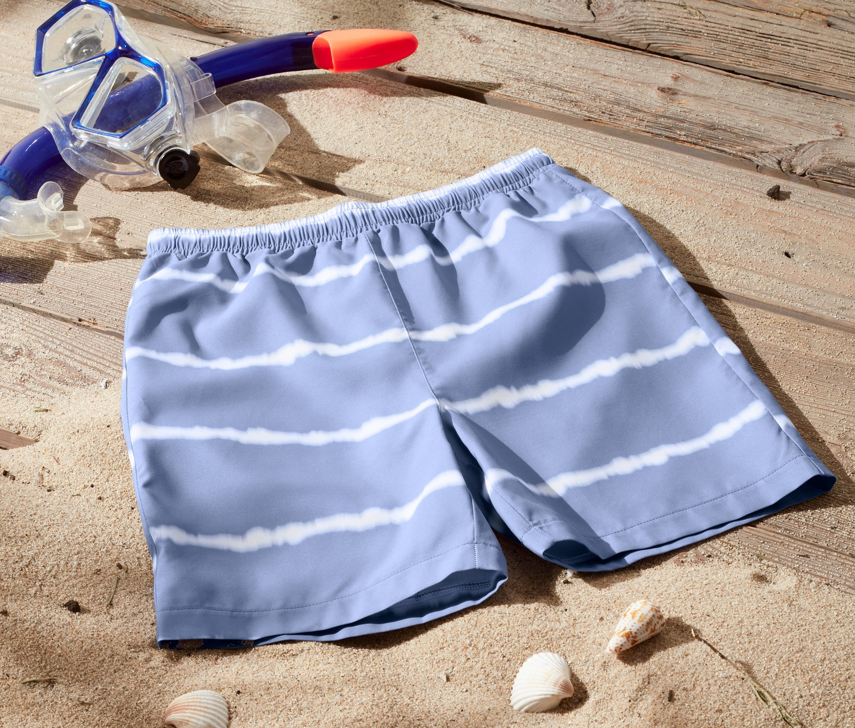 Badeshorts aus recyceltem Material liegen im Sand neben Muscheln, Schnorchel und Surfbrett.