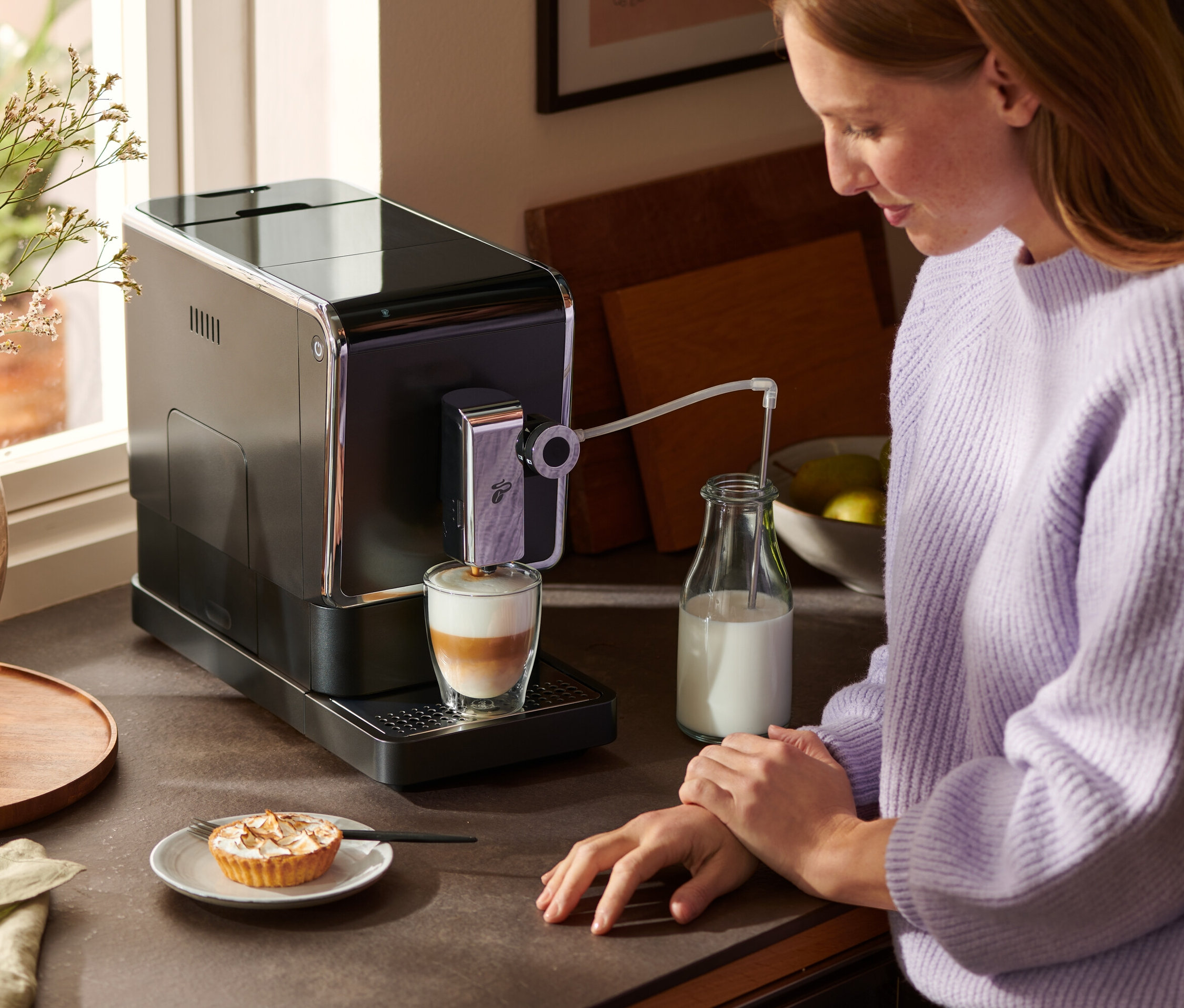 Eine Frau steht an einer Küchentheke und bereitet mit einem anthrazitfarbenen »Esperto Pro« Tchibo Kaffeevollautomaten einen Latte zu. Neben dem Kaffeevollautomaten stehen eine Milchflasche und ein Teller mit Kuchen.