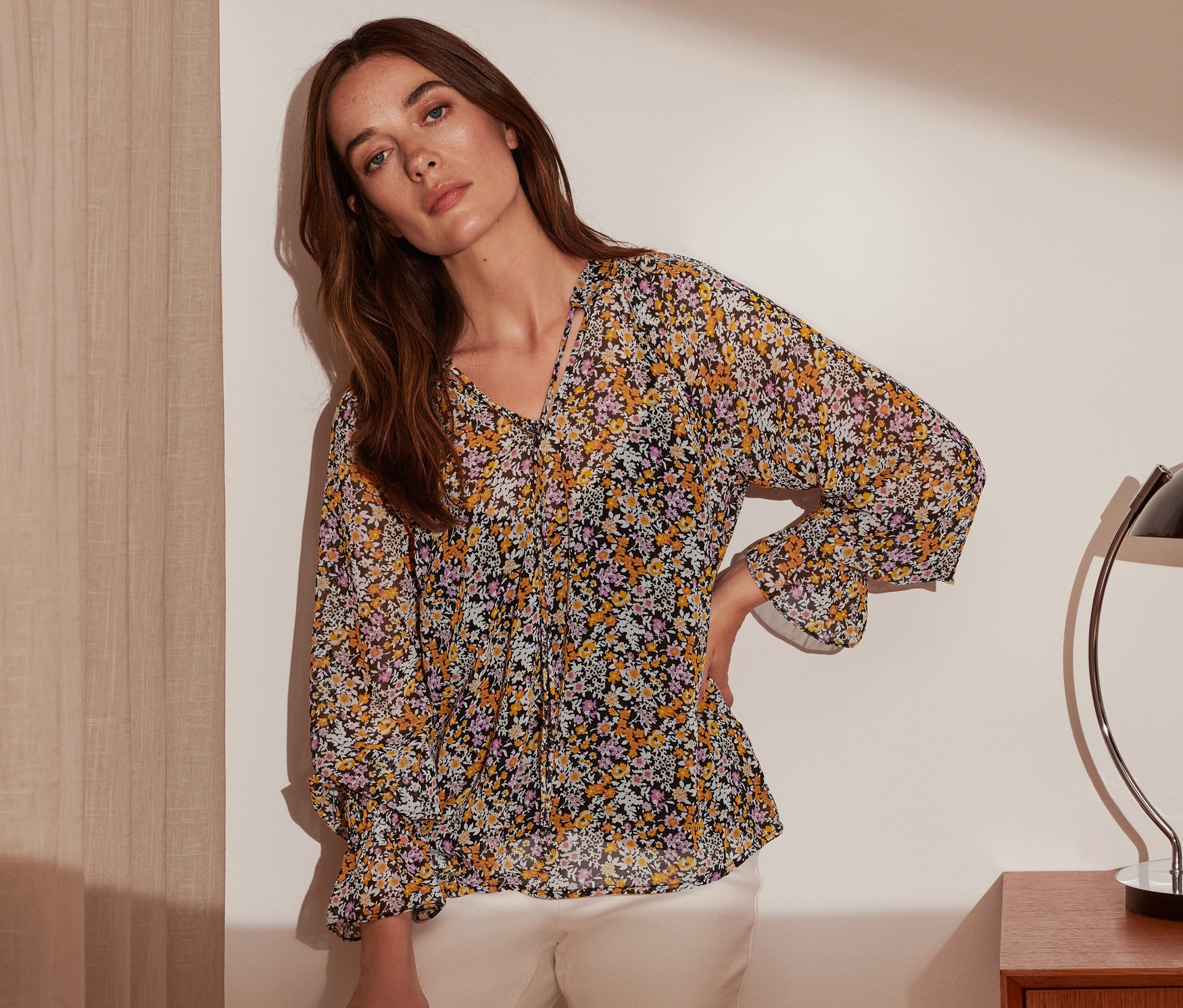 Eine Frau posiert in einer Bluse mit Print und weißen Hosen.