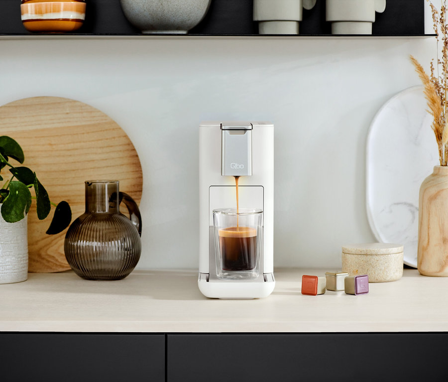 Qbo ICONIC Dark Chrome und Qbo ESSENTIAL Bright Sand Kaffeemaschine füllt ein Glas mit Kaffee. Daneben stehen Kapseln.