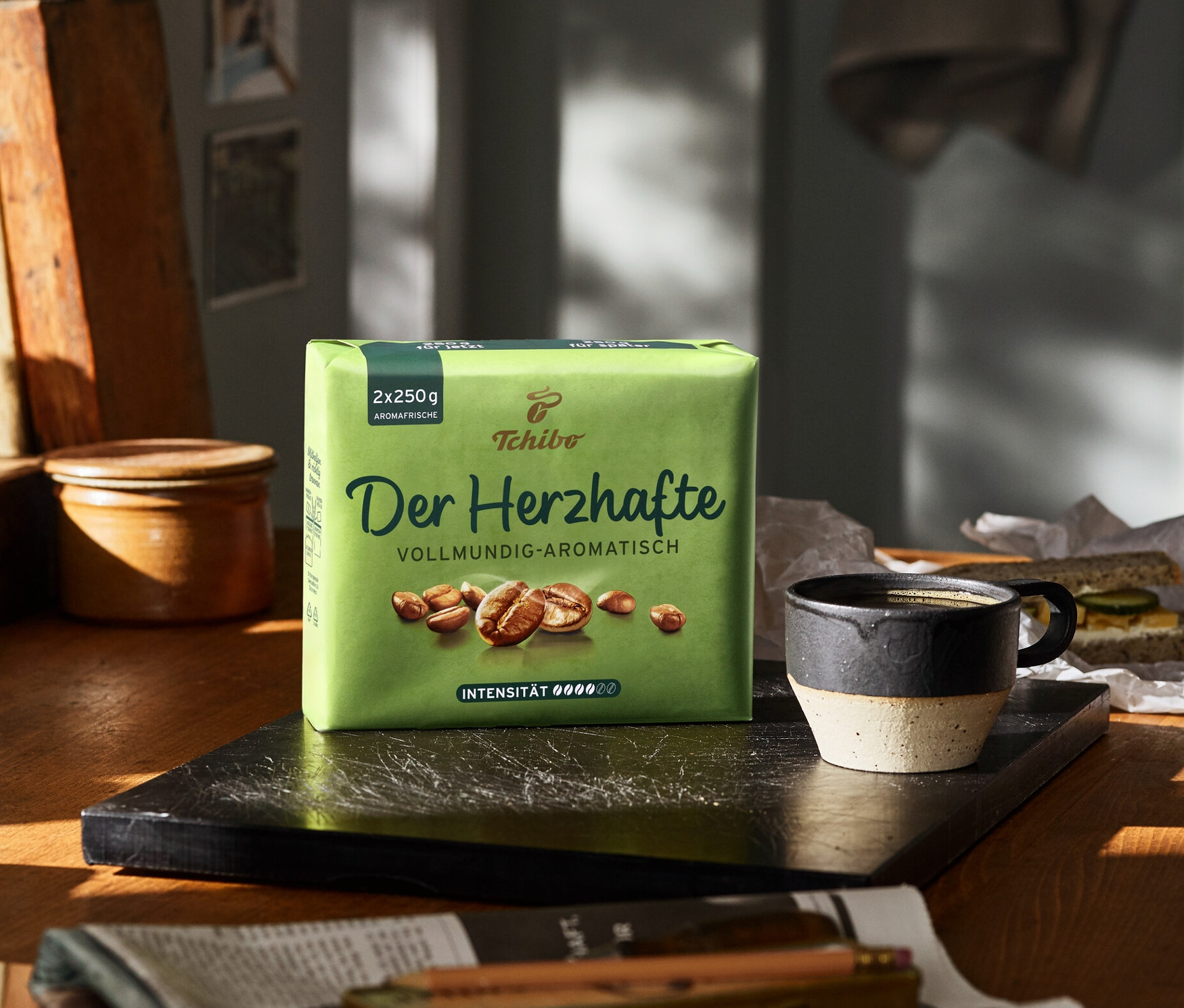 Auf einem Tisch steht eine Packung Der Herzhafte - 500 g Gemahlen Kaffee neben einer Tasse und einem Sandwich.