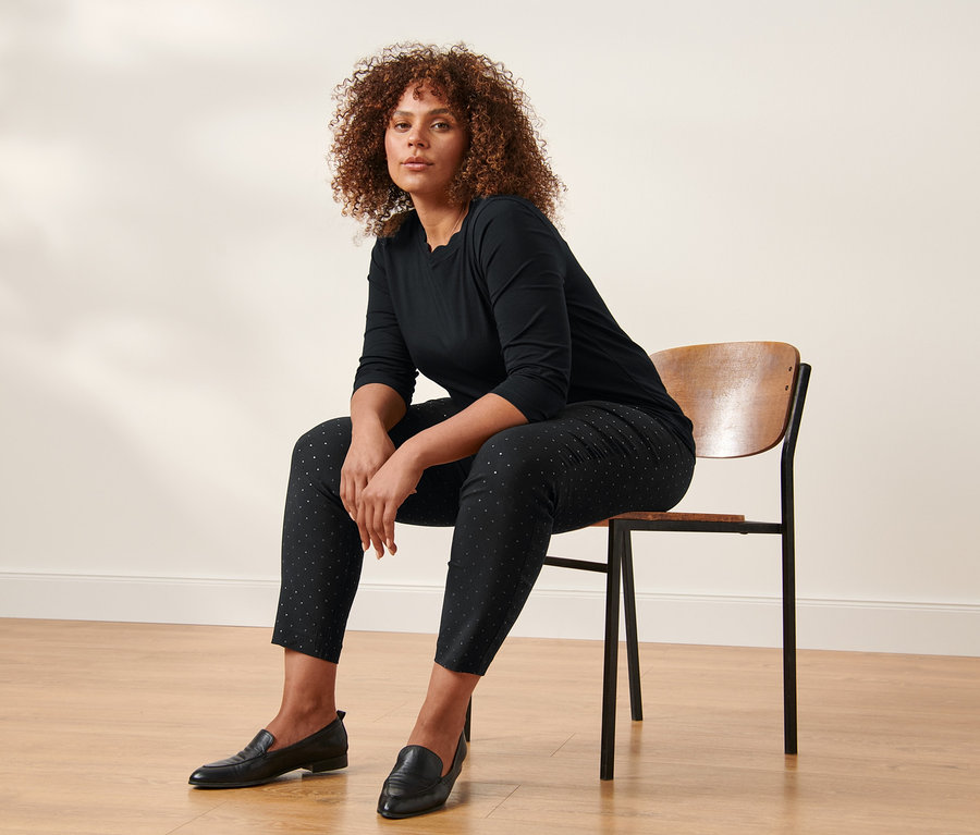 Eine Frau sitzt auf einem Stuhl und trägt ein schwarzes Shirt mit 3/4-Arm und eine gepunktete Stretchhose.