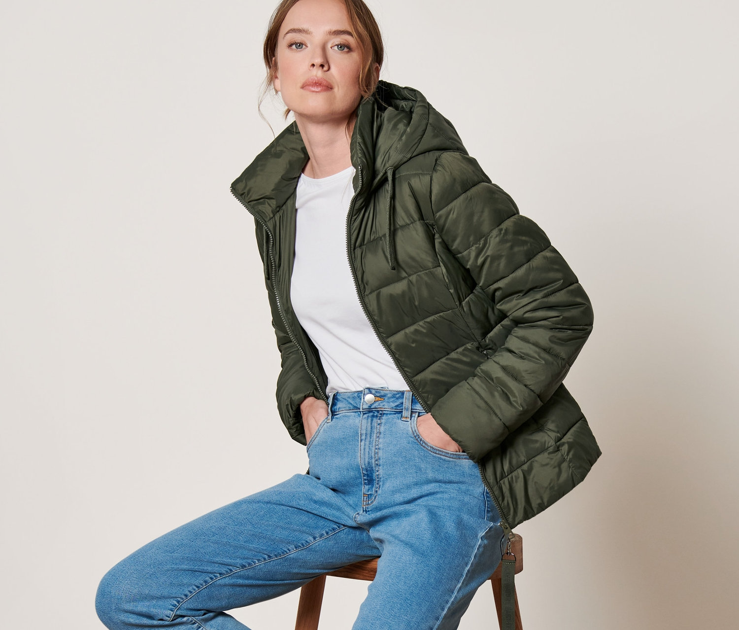 Frau sitzt auf Hocker, trägt olivgrüne Steppjacke mit Kapuze.