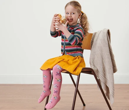 Mädchen sitzt auf einem Stuhl und isst einen Doughnut. Sie trägt einen gelben Rock und eine rosa 5 Kinder-Baumwoll-Strumpfhosen.