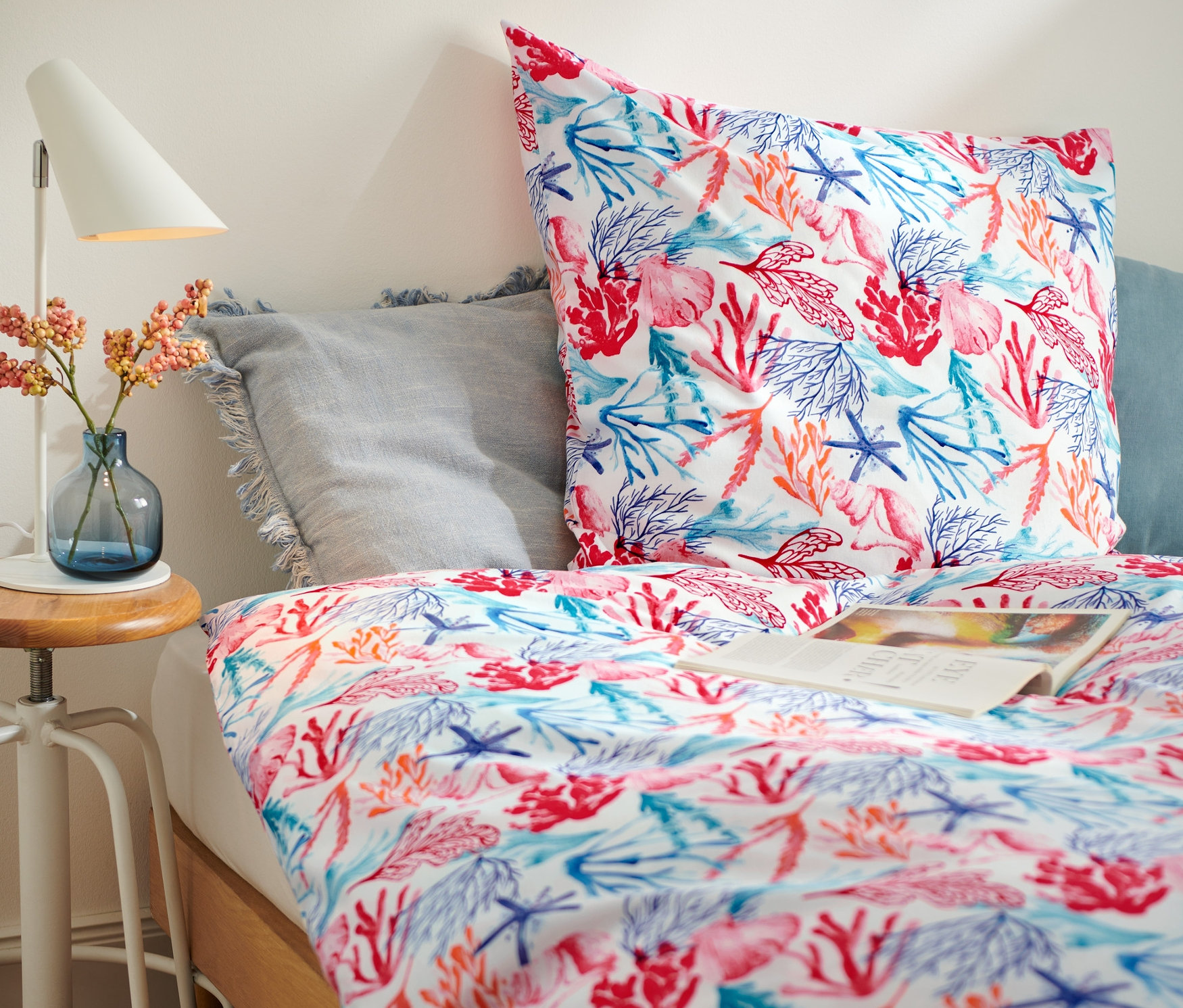 Auf einem Bett liegen drei Renforcé-Bettwäsche-Sets in Normalgröße mit Meeresmotiven. Neben dem Bett steht ein Tisch mit Lampe und Vase.