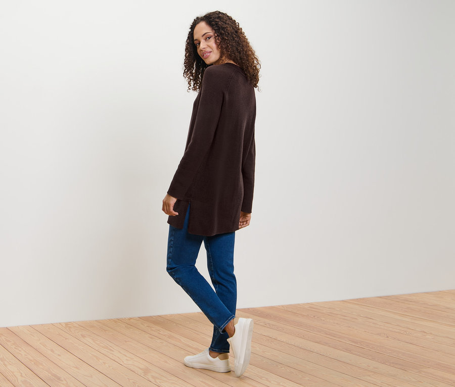 Frau blickt über die Schulter und posiert in einem langen, dunkelbraunen Strickpullover, Jeans und Turnschuhen.