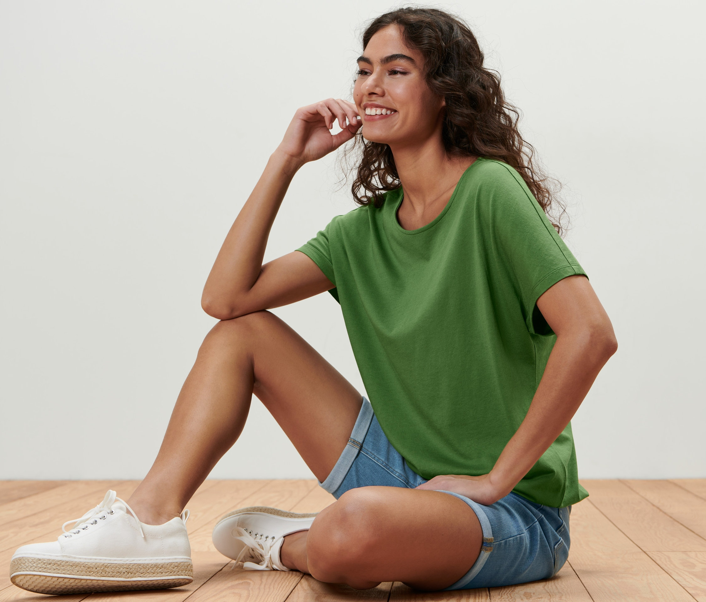 Frau sitzt auf dem Boden und trägt ein grünes Blusenshirt, Jeansshorts und weiße Turnschuhe.