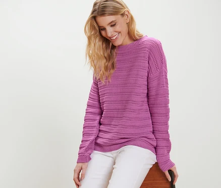 Blonde Frau sitzt und trägt einen lila Pullover mit Ajour-Muster und weiße Hose.