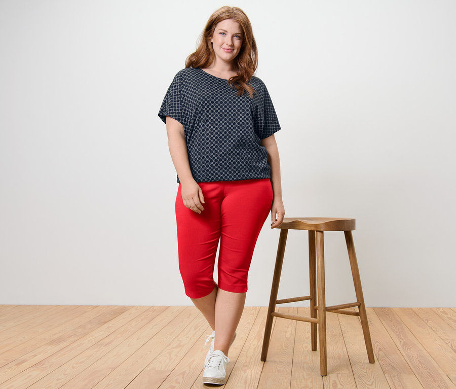 Eine Frau steht neben einem Holzhocker und trägt ein bedrucktes Blusenshirt und eine rote 3/4-Stretchhose.