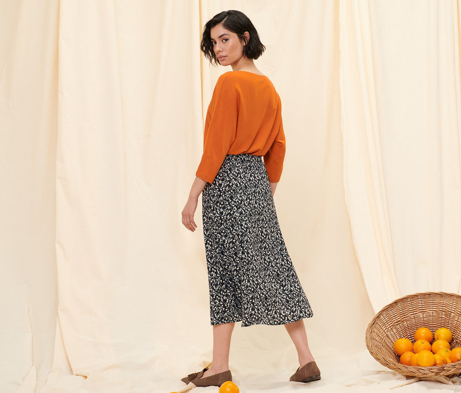 Frau posiert in orangefarbenem Feinstrickpullover, floralem Midi-Rock und Loafern aus Veloursleder.
