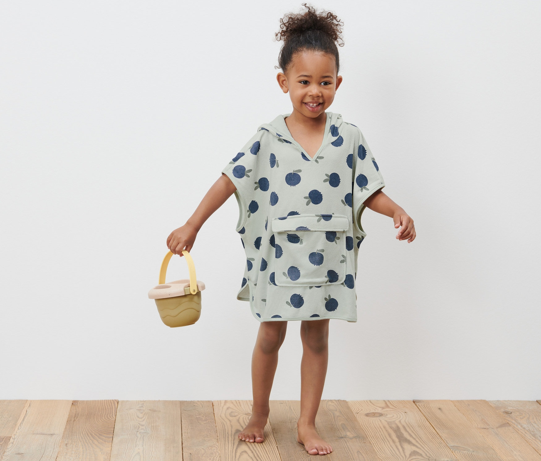 Lächelndes Mädchen trägt einen Kleinkind-Frottierponcho und hält einen gelben Eimer.