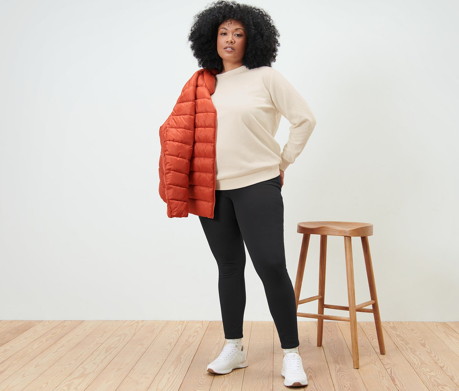 Frau mit Afro steht mit einer orangefarbenen Jacke über der Schulter und einem Sweatshirt mit Stickerei.