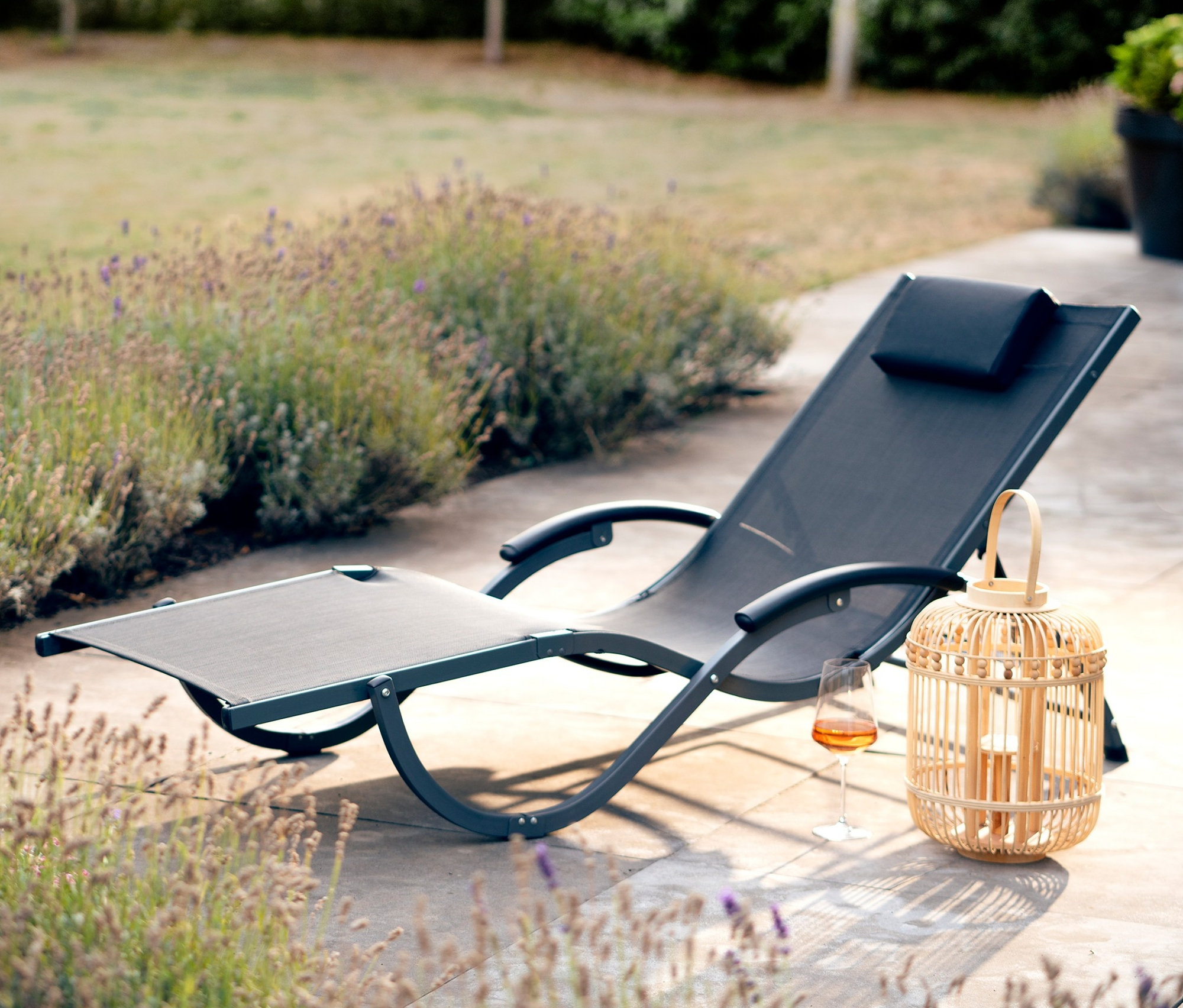 Eine schwarze Leco Sonnenliege steht auf einer Terrasse neben einem Weinglas und einer Laterne.