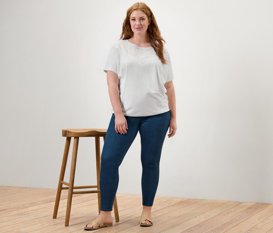 Frau steht und trägt ein weißes Blusenshirt mit Stickerei und Jeggings.