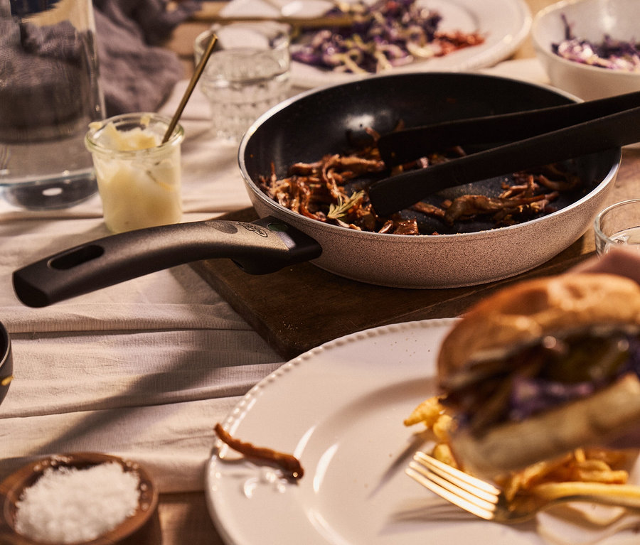 Auf einem gedeckten Tisch steht eine BALLARINI Aluminium-Bratpfanne (sand, 24 cm) mit Speisen, daneben ein Wasserglas und im Vordergrund hält eine Hand einen Burger.