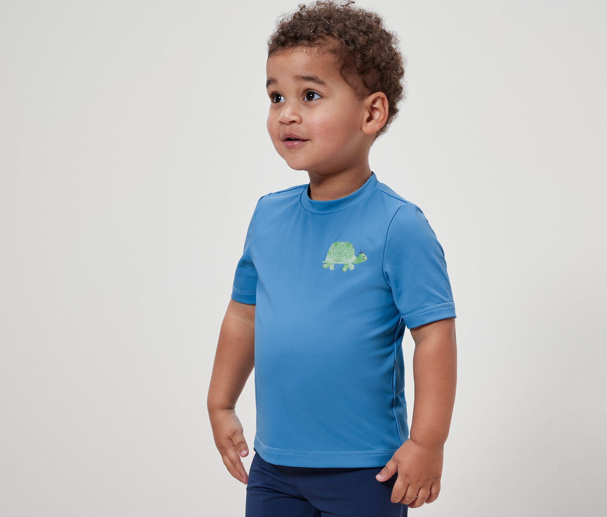 Ein Junge trägt ein blaues Kinder-Shirt mit UV-Schutzfaktor 80 und eine dunkelblaue Hose.