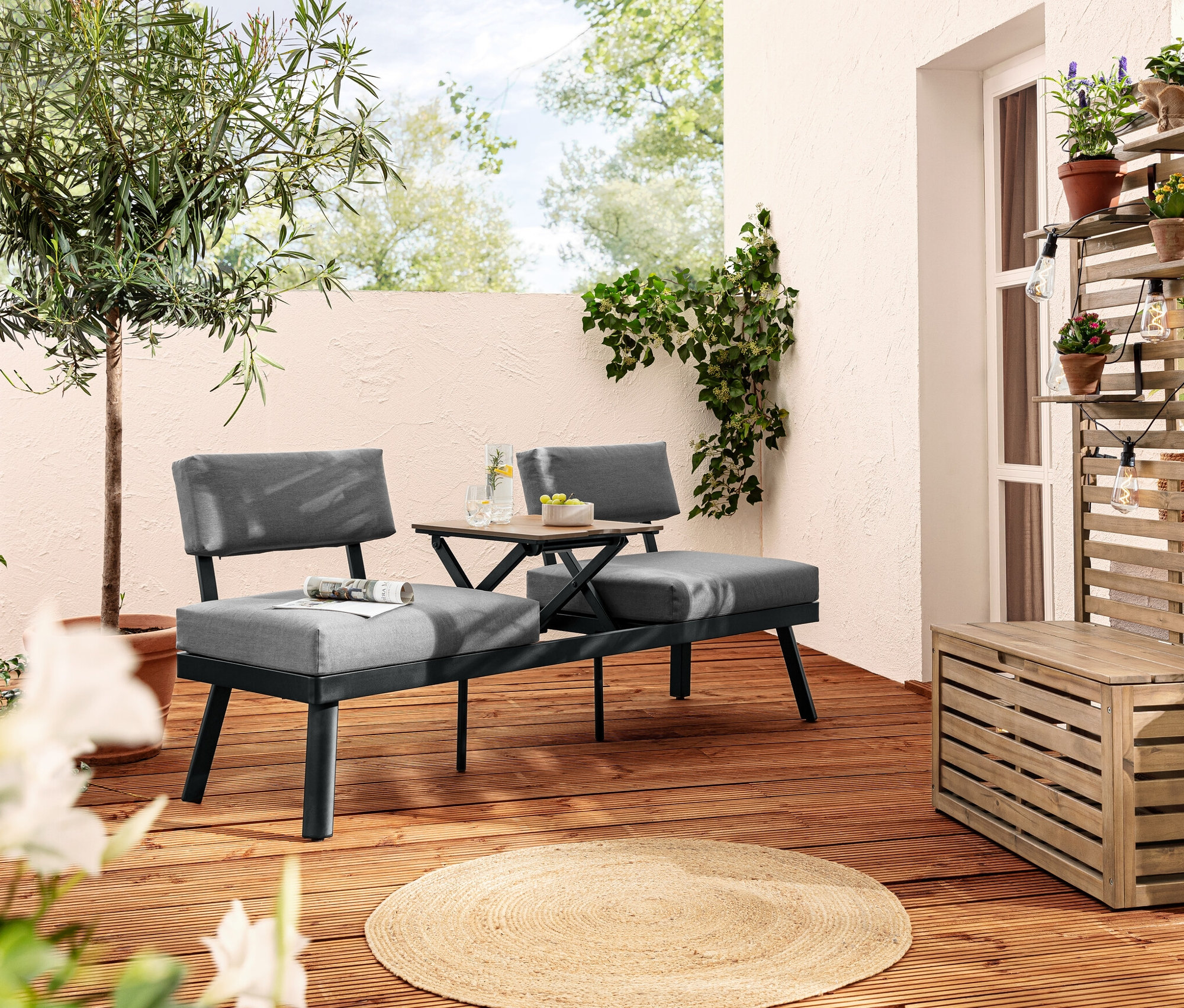 Zwei graue 2-in-1-Sofa-Liegen mit hochklappbarem Mitteltisch stehen auf einer Holzterrasse. Eine LED-Filament-Lichterkette hängt an einer Holzkonstruktion mit Blumen.